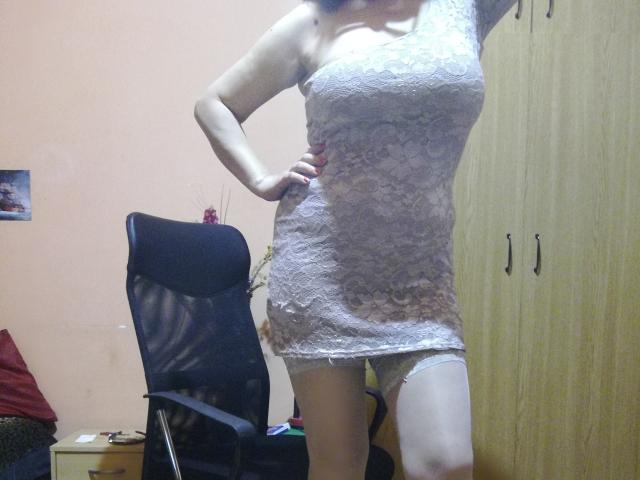 LadyEvelynn trägt ein weißes Spitzenkleid, das ihre Brüste zeigt, mit einer Hand auf der Hüfte, und steht in einem Raum mit einem Stuhl und einem Holzschrank..