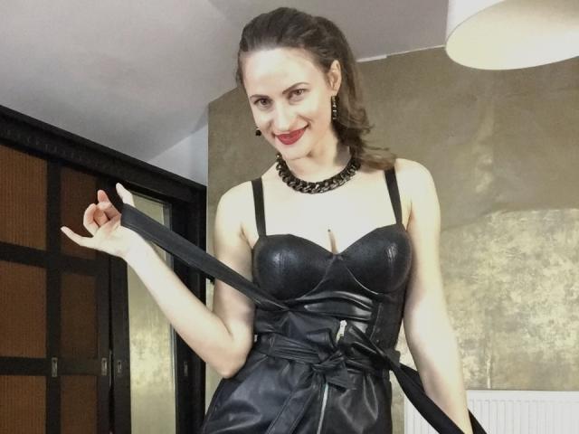 AbyX has pale skin, brown hair in a ponytail, red lipstick, black leather dress with straps, cleavage, black choker, holding dress strap, smiling, in a room.