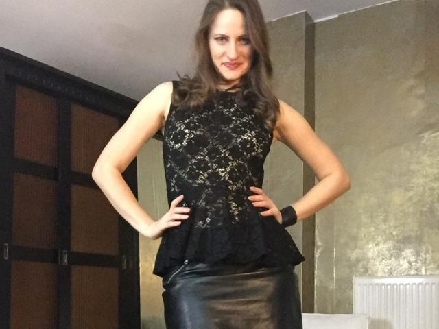 AbyX with hands on hips, wearing a black lace sleeveless top and a black leather skirt, smiling, has wavy brown hair, and a black wristband.