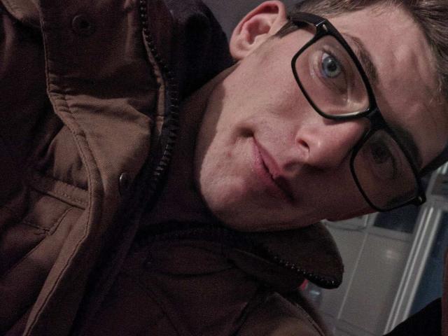 Colin Basson, avec des lunettes, une veste marron et un léger sourire, des yeux bleus, une peau claire, des cheveux bruns courts, un anneau au nez, un selfie en gros plan, la tête légèrement inclinée, regardant la caméra..