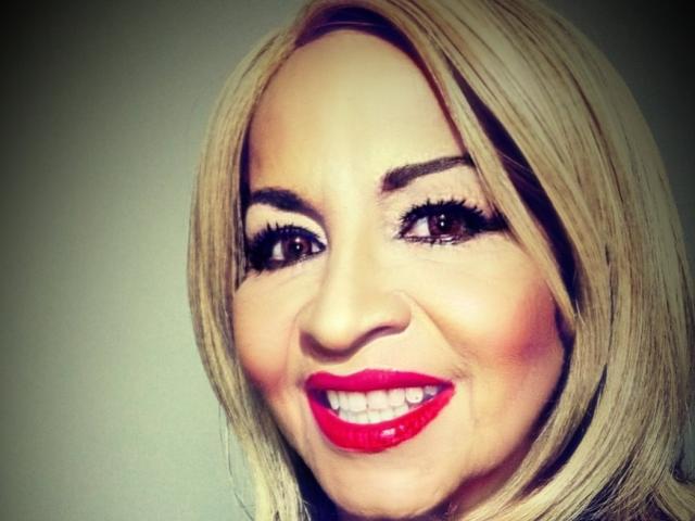 SexyLilith tiene el cabello rubio, lápiz labial rojo y una sonrisa radiante. Sus grandes ojos marrones están enmarcados por cejas gruesas y oscuras. Está mirando directamente a la cámara..