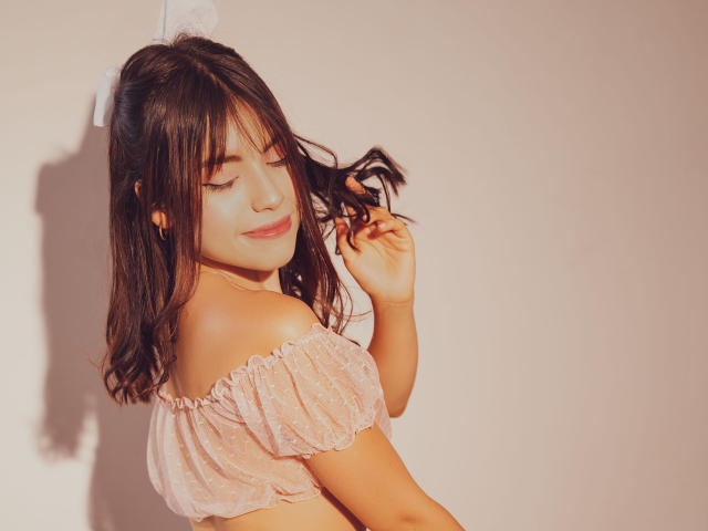 VanityValos with medium tits, wearing a pink, off-shoulder, sheer top, has dark brown hair, and a white bow, smiling with eyes closed, holding her hair.
