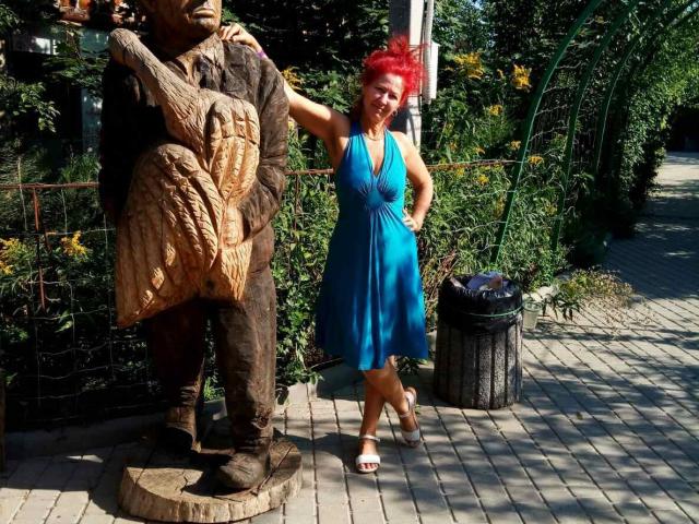 MilayaDFlow mit leuchtend roten Haaren, die ein blaues Kleid trägt, steht neben einer hölzernen Statue mit einer großen, geschnitzten, fellartigen Brust. Sie lächelt und berührt den Arm der Statue..