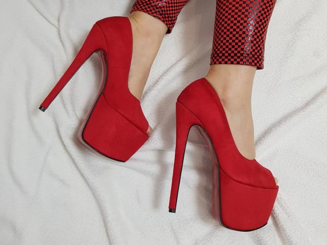 CruelDomme's red, high-platform, peep-toe heels with long, narrow stiletto heels and red checkered, shiny pants, showing off her bare, pale, slender feet on a white surface.