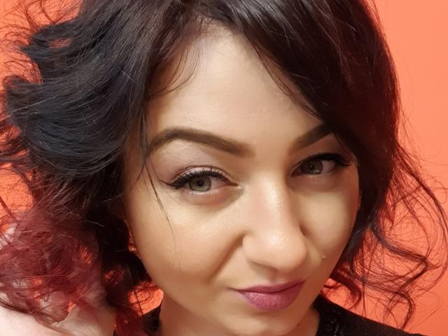 JolieXKitty has wavy black hair, light brown eyes with long eyelashes, and a subtle pink lip. She's smiling with a slightly upturned nose and a smooth, fair complexion.