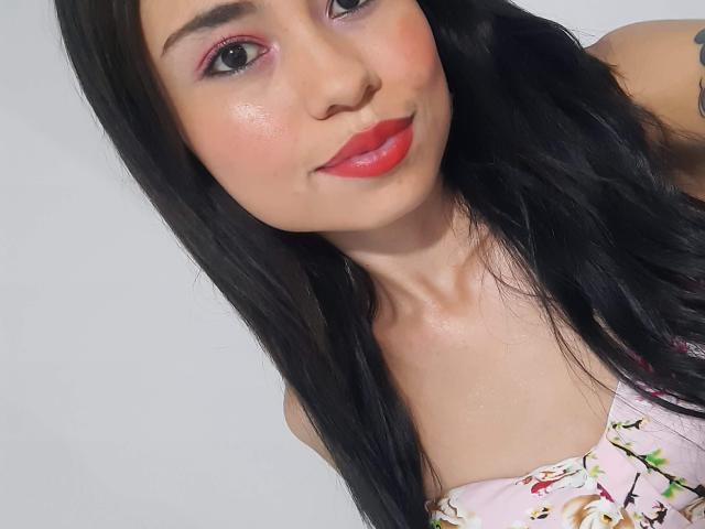 LolyFunX ha una pelle luminosa, rossetto rosso e capelli neri, indossa una maglietta rosa con fiori e i suoi seni sono appena visibili. Sorride con l'ombretto rosa e un viso carino..