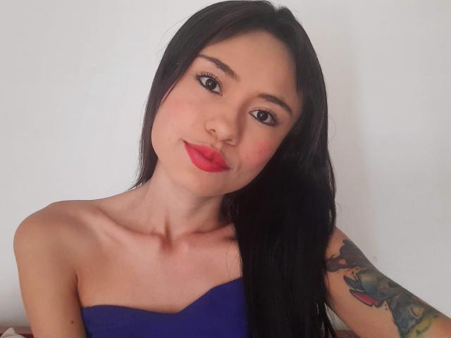 LolyFunX mit langen schwarzen Haaren, rotem Lippenstift und einem blauen Top ohne Träger, das einen tätowierten Arm zeigt, hat ein süßes Gesicht und kleine, pralle Brüste..
