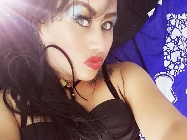 CathyCat com cabelo preto, sombra azul, batom vermelho, sutiã preto, tatuagem no ombro direito, colar preto, olhando para a câmera, luz do sol no lado esquerdo, fundo azul e branco..