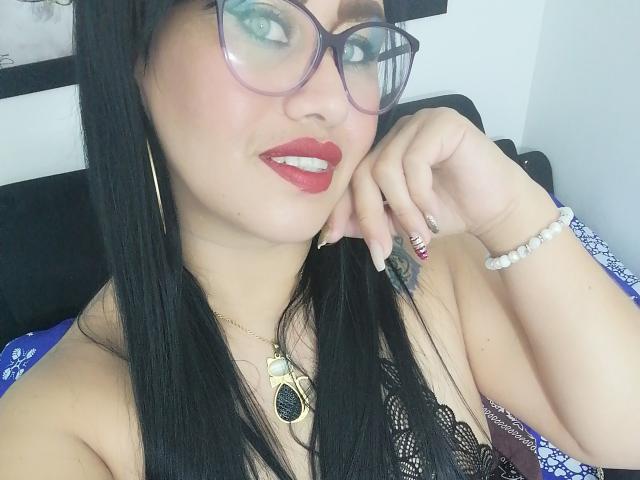 CathyCat, com cabelo preto, batom vermelho, óculos e um colar, exibindo seus seios em uma blusa de renda, usando uma pulseira de pérolas e com as unhas listradas..