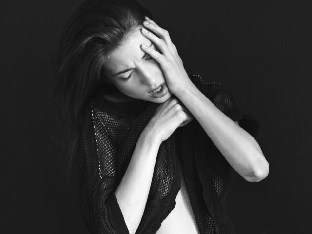 StuartKate with long hair, eyes closed, biting lower lip, wearing a black mesh top, hands touching head, black and white, looking pained or stressed, soft skin, delicate features.