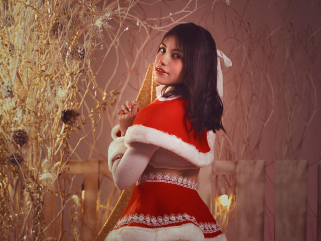VanityValos, con la pelle marrone media, i capelli scuri e ondulati e un sorriso carino, indossa un costume rosso da Babbo Natale con pelliccia bianca, guarda oltre la spalla e tiene in mano un nastro dorato..