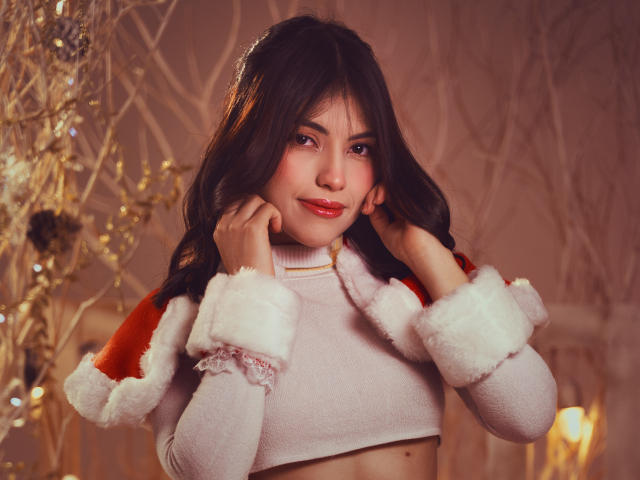 VanityValos con capelli scuri e ondulati, un cappotto rosso da Babbo Natale, polsini di pelliccia bianca, un top corto rosa e rossetto rosso, si tiene le guance, sorridendo, in un ambiente caldo e con una luce soffusa..