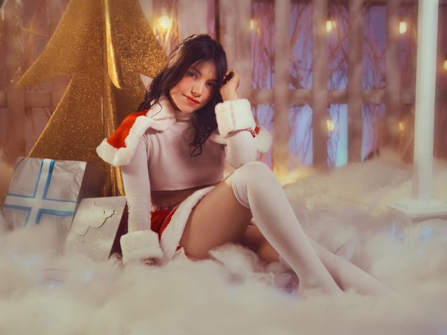 VanityValos, com cabelos escuros e ondulados, vestindo um capuz vermelho de Papai Noel, meias brancas até a coxa e uma blusa cropped branca, sentada em "neve" branca e fofa, com uma árvore de Natal dourada..