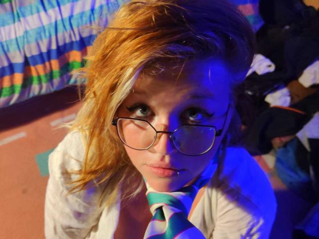 Shushaina, avec ses cheveux blonds en bataille, ses lunettes et son piercing au lobe, portant une cravate rayée et une chemise blanche, regardant vers le haut avec un éclairage bleu et orange sur son visage..