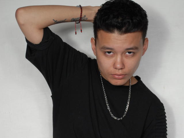 EmiFTM, avec des cheveux noirs courts, portant une chemise noire, un collier en chaîne et un bracelet de perles rouges, a un tatouage sur l'avant-bras droit et une expression neutre..