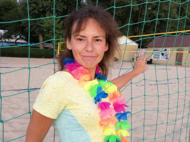 GrostFlow avec de petites seins, portant une chemise jaune, un collier de fleurs colorées et des cheveux bruns ondulés, faisant un signe de la paix devant un filet vert. Souriante, dans un décor de plage..