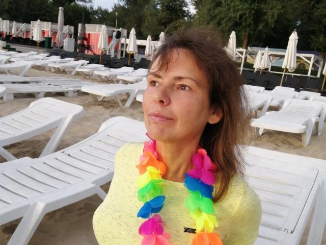 GrostFlow, con i capelli castani, indossa una camicia gialla e una ghirlanda colorata, ha un sorriso leggero e mostra delle piccole lentiggini sul viso, ed è in piedi di fronte a delle poltrone bianche..