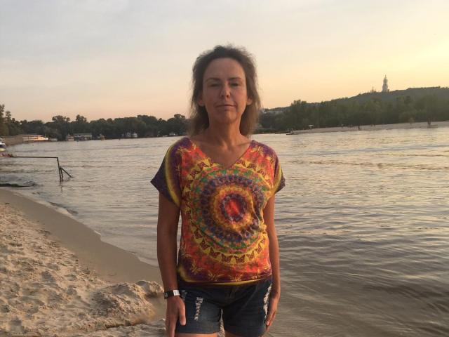 GrostFlow stands on a beach, wearing a colorful tie-dye shirt and blue jeans, with sandy feet and a wristwatch, against a calm river and sunset.