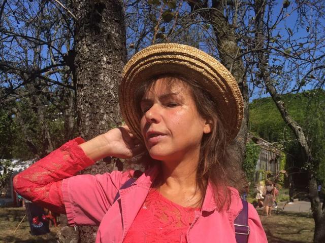 GrostFlow with brown hair, wearing a straw hat, pink lace shirt, and pink jacket, holding her hat, showing a slightly sunburned face, and a relaxed expression.