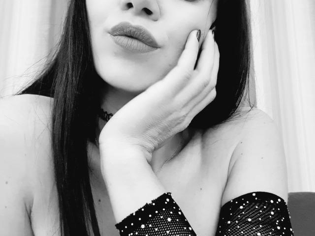 Il viso di SparklyEve, con capelli neri e lisci, indossa una manica trasparente con paillettes, una mano sul viso, una collana nera e labbra piene, in una foto in bianco e nero..