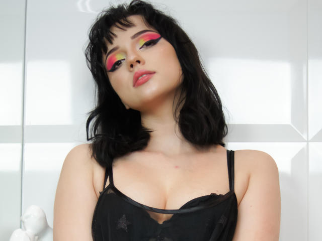 DeniseKitty with black hair, pink and yellow eye makeup, red lips, black lace bra, and small tits, leaning slightly forward, looking up with a confident expression.