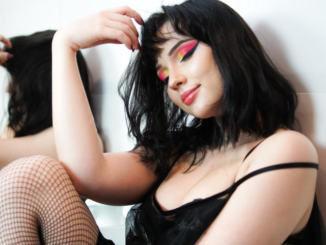 DeniseKitty, avec ses cheveux noirs, son maquillage oculaire rose et jaune, sa lingerie noire et ses bas en résille, assise avec une main dans ses cheveux, montrant ses seins et un léger sourire..