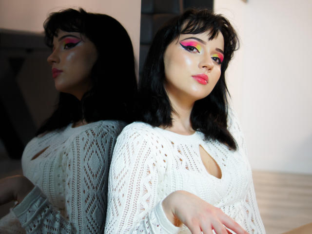 DeniseKitty with black hair, white knit top, pink eyeshadow, yellow eyeliner, and pink lipstick, leaning against mirror showing reflection, showcasing her shiny skin and subtle smile.
