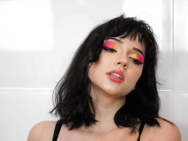 DeniseKitty, con cabello negro, maquillaje de ojos rosa y amarillo, y labios rosados brillantes, ligeramente babeando, vestida con una blusa negra, tiene una expresión sexy y seductora, y piel pálida..