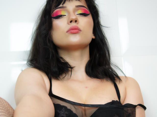 DeniseKitty with black lace bra, pink and yellow eye makeup, pouty pink lips, black wavy hair, white tiled background, holding selfie, sexy, confident, showing off her tits.