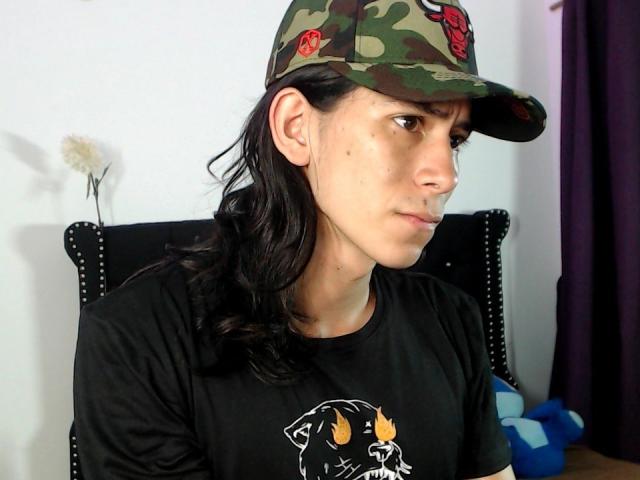 Antonio Santos, con cabello negro largo, usando una gorra de camuflaje con flores rojas y una camiseta negra con un perro de dibujos animados, mirando hacia un lado con una expresión seria..