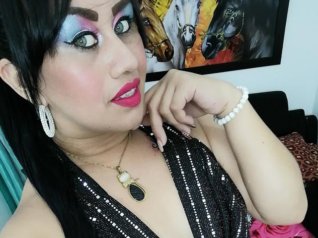 CathyCat has pink lipstick, black hair, and wears a black sequined dress with a gold pendant. She's touching her chin, showing off white earrings and a bead bracelet.