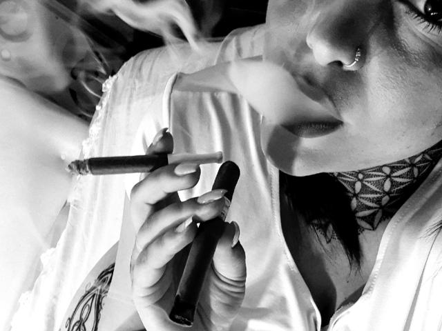 "SmokingGoddess" con un piercing en la nariz, sosteniendo un cigarrillo en una mano y un tubo negro en la otra, vistiendo una camisa blanca con tatuajes visibles. Blanco y negro..