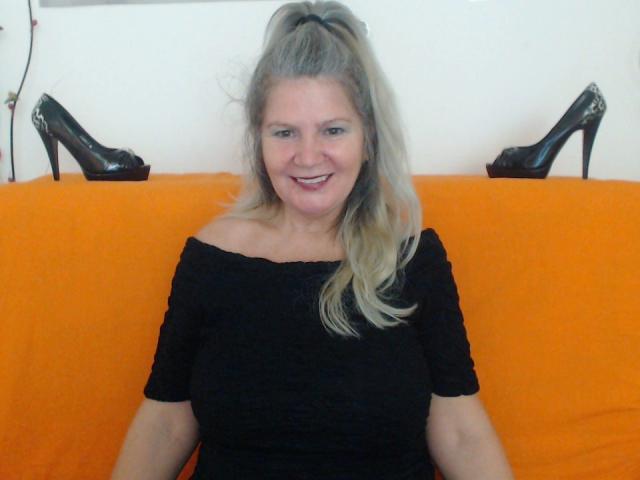 AdamaXbella with blonde ponytail, smiling, wearing black off-shoulder top, sitting on orange couch, black high heels on white wall behind her. Boobs visible under top.