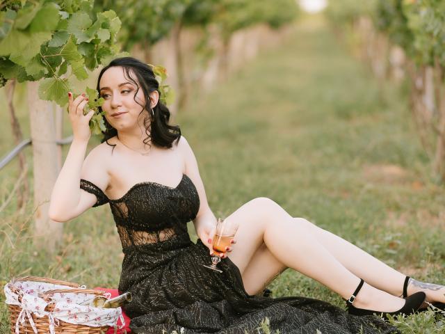 ClaptonFow, with wavy black hair, wearing a black lace dress, sitting in a vineyard, holding a glass of wine, touching leaves, legs crossed, black heels, wicker basket.