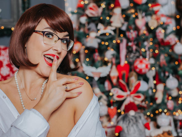 CaroJones with short brown bob, glasses, red lipstick, white shirt off-shoulder, pearl necklace, finger to lips, Christmas tree with ornaments in blurred background, looking surprised.