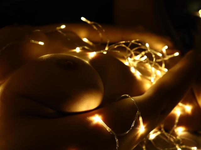 LailaNoire's tits glowing with warm fairy lights, her nipples clearly visible, lying down in soft, dark lighting. Her skin is smooth and the lights create gentle, sparkling highlights.