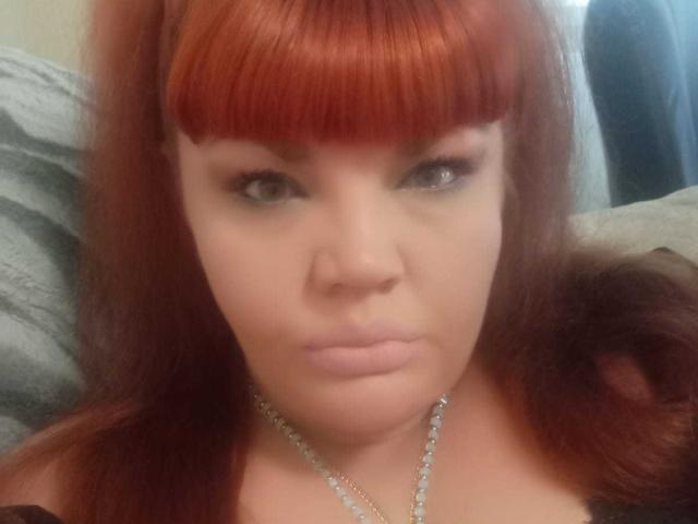 RedBustyX has red hair with blunt bangs, pale skin, and a serious expression. She's wearing a black top and a silver necklace, with her tits slightly visible.