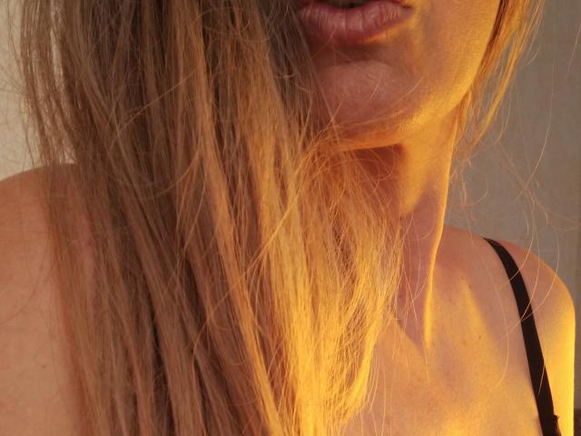 Mayacorse's face with long, messy blonde hair partially covering her lips, sunlit skin, black strap, and visible neck. Her lips are slightly parted, and she's wearing a black top.