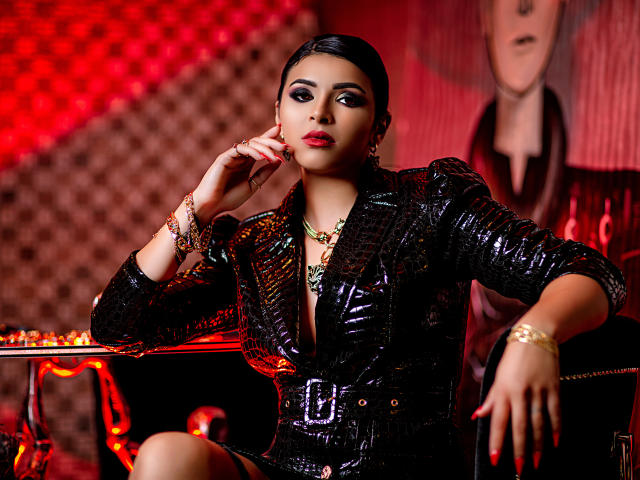 Devasha with shiny black leather jacket, red lipstick, and gold jewelry, sitting confidently, hand near face, looking sexy with her ass slightly raised, in a red-lit, stylish setting.