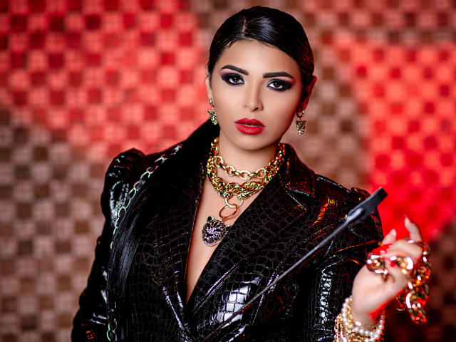 Devasha with dark hair, red lipstick, and gold jewelry, wearing a black, shiny, croc-pattern blazer, holding a black whip, looking fierce, with a gold chain necklace.
