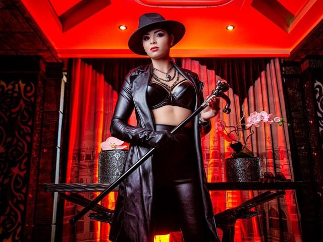 Devasha with black hat, leather coat, and gloves holding a whip, wearing a black leather bra, and tight leather pants, standing in a red-lit room with ornate decor.
