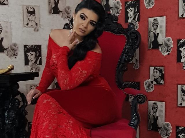 Devasha with black hair in a red lace off-shoulder dress, sitting on an ornate red chair, her hand touching her neck, looking seductive with her ass prominently highlighted.