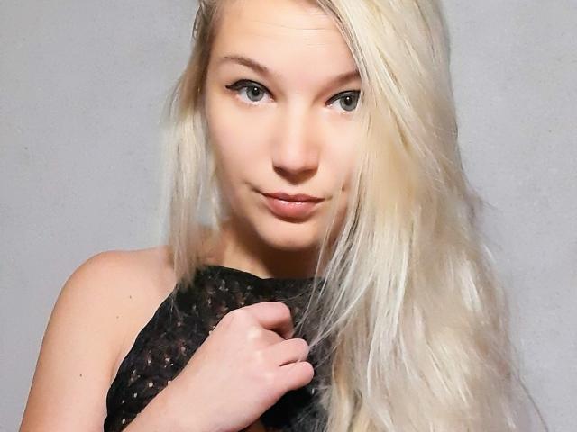 AlmaDeAngel69 mit langen blonden Haaren, heller Haut und dezenter Schminke, trägt ein schwarzes, durchsichtiges Oberteil, das sie mit ihrer rechten Hand hält, und blickt mit einem leichten Lächeln in die Kamera..