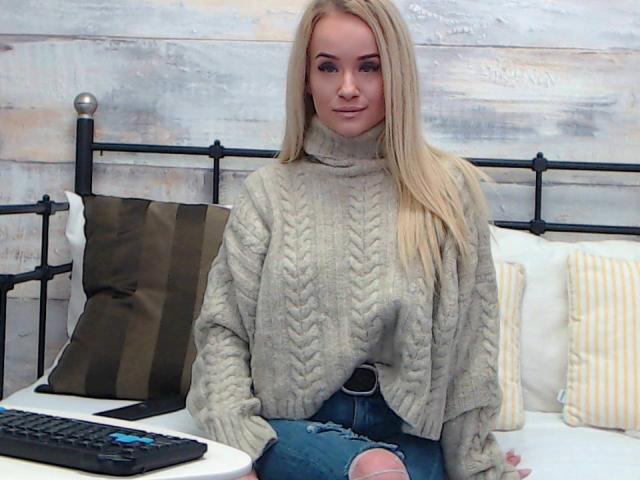 SophieXDee with long blonde hair, wearing a chunky gray knit sweater and ripped blue jeans, sitting on a bed with a black metal headboard, and a keyboard.