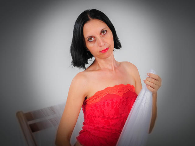SexyFlora with short black hair, red lace strapless dress, holding white fabric, smiling, looking at camera, fair skin, red lipstick, small breasts, wooden chair partially visible.