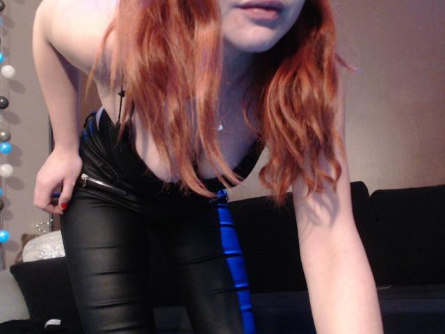 LilouLove with red hair, leaning forward in black leather pants and blue tie, showing cleavage and arms. She's wearing a necklace and has pale skin.