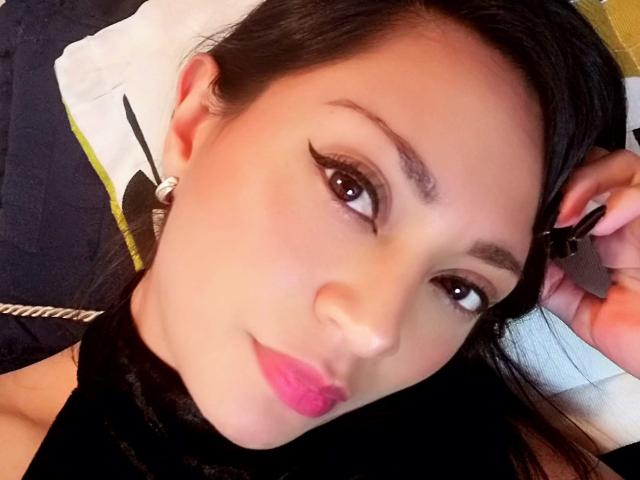 DakotaSweet, avec ses cheveux noirs, ses yeux marrons et son rouge à lèvres rose, tenant une pince à cheveux noire, portant des boucles d'oreilles noires et blanches, regardant l'appareil photo avec un léger sourire..