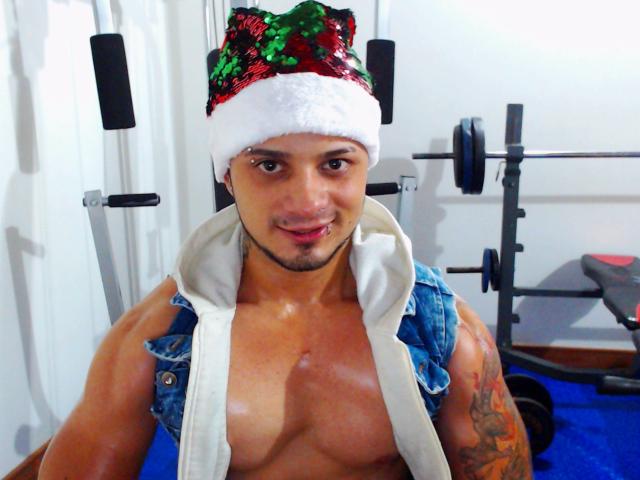 Everet draagt een rode en groene kerstmuts met pailletten, een vest van denim zonder mouwen, een witte handdoek, en geen shirt, met een tatoeage op zijn rechterarm, en glimlacht in een sportschool..
