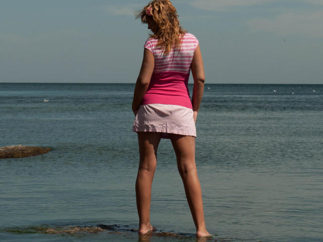 AdamaXbella stands barefoot on rocky shore, facing ocean, with wavy blonde hair, pink and white striped shirt, pink skirt, showing her ass, in sunlit water, legs apart.