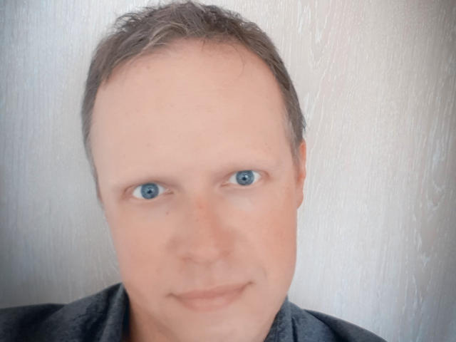 NaughtyHugues tiene un rostro pálido, ojos azules, cabello corto y castaño, y una ligera sonrisa. Lleva una camisa gris. La imagen está ligeramente borrosa..